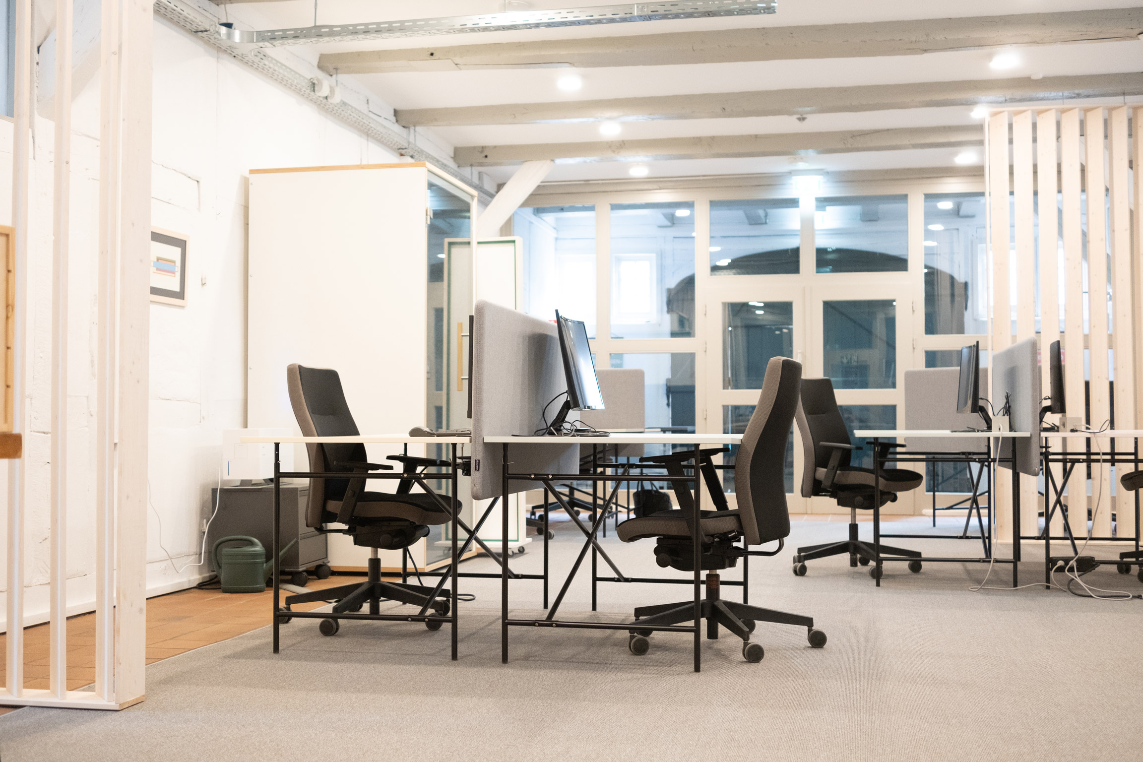 Helle, moderne Coworking-Arbeitsplätze mit Schreibtischen, Bürostühlen und Monitoren in einem offenen Raum mit Deckenbalken.