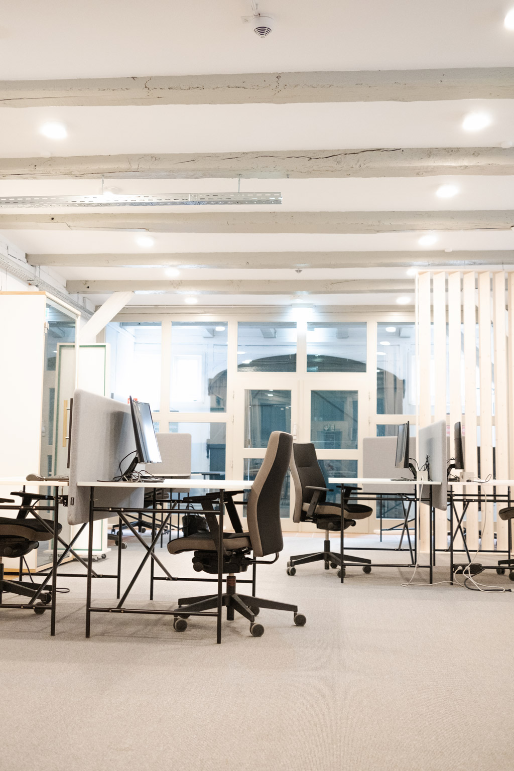 Helle, moderne Coworking-Arbeitsplätze mit Schreibtischen, Bürostühlen und Monitoren in einem offenen Raum mit Deckenbalken.