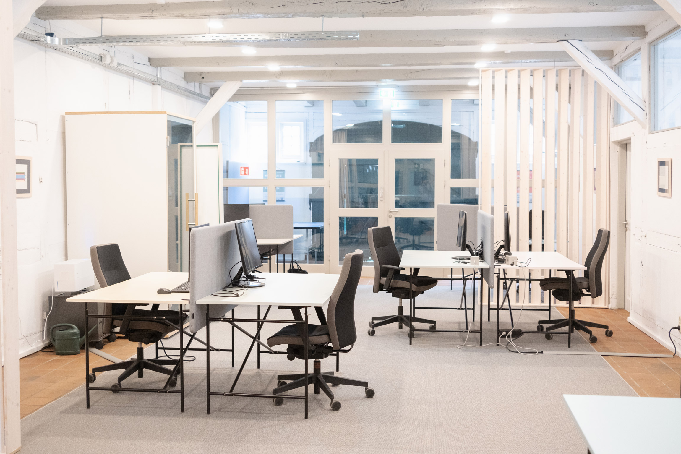 Helle, moderne Coworking-Arbeitsplätze mit Schreibtischen, Bürostühlen und Monitoren in einem offenen Raum mit Deckenbalken.